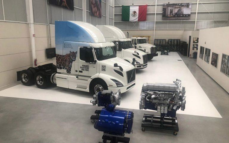 Line of white semi-trailers in a spacious garage, foreground showing two large engines on stands and a blue-wrapped truck with a leopard graphic at the front.