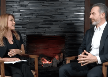 Woman host interviewing a man in a studio, seated in wooden chairs beside a lit stone fireplace.