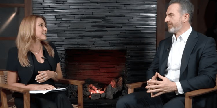 Woman host interviewing a man in a studio, seated in wooden chairs beside a lit stone fireplace.