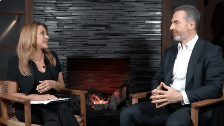Woman host interviewing a man in a studio, seated in wooden chairs beside a lit stone fireplace.
