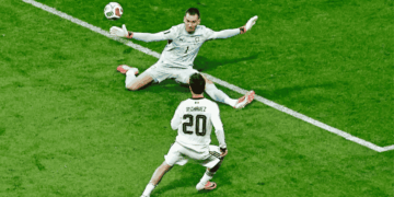 Soccer goalkeeper dives to save the ball as an opposing player wearing number 20, M. Chavez, approaches on a green pitch.