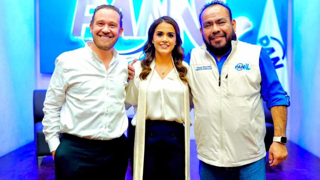 Three adults pose together in a bright room with a blue neon sign in the background. Left man in white shirt, center woman in cream cardigan, right man in PANL vest.