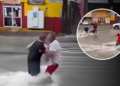 People wading through knee-deep floodwater on a city street, one man in a white shirt helping another in red shorts; storefronts visible in the background.