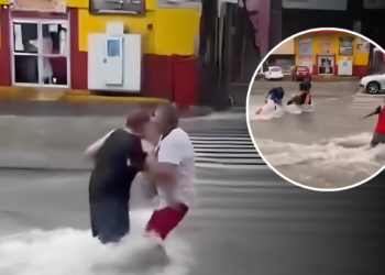 People wading through knee-deep floodwater on a city street, one man in a white shirt helping another in red shorts; storefronts visible in the background.