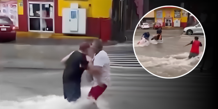 People wading through knee-deep floodwater on a city street, one man in a white shirt helping another in red shorts; storefronts visible in the background.