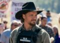 Man in a black cowboy hat and tactical vest labeled 'U.S. Marshal' stands at a protest, looking to the side.
