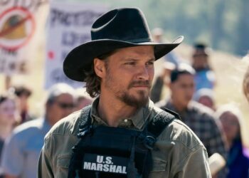 Man in a black cowboy hat and tactical vest labeled 'U.S. Marshal' stands at a protest, looking to the side.