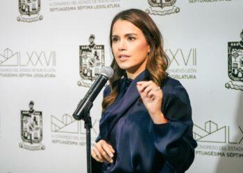 Woman in a navy blouse speaks into a handheld microphone at a press conference, with government insignias on the backdrop behind her.