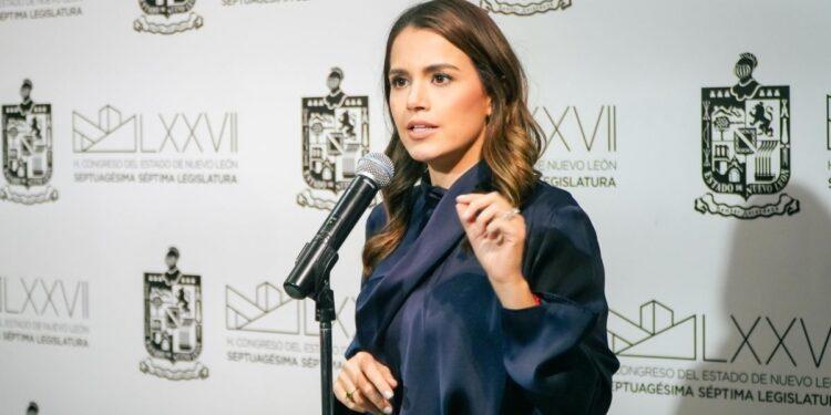 Woman in a navy blouse speaks into a handheld microphone at a press conference, with government insignias on the backdrop behind her.